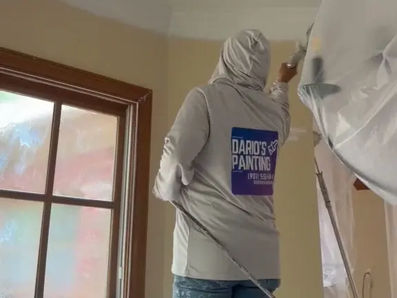 A Dario’s Painting crew member in a branded hoodie spraying a ceiling, with the window and walls masked in plastic
