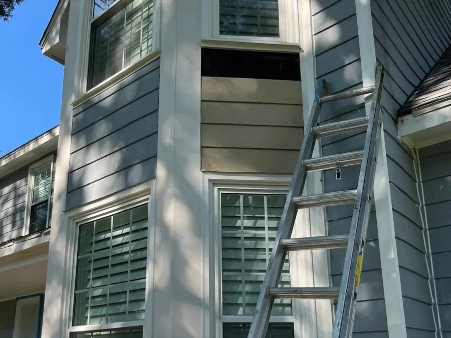 Tan lap siding set between two windows on a blue-grey house, with a dark gap left open above it and an aluminum ladder leaning on the wall