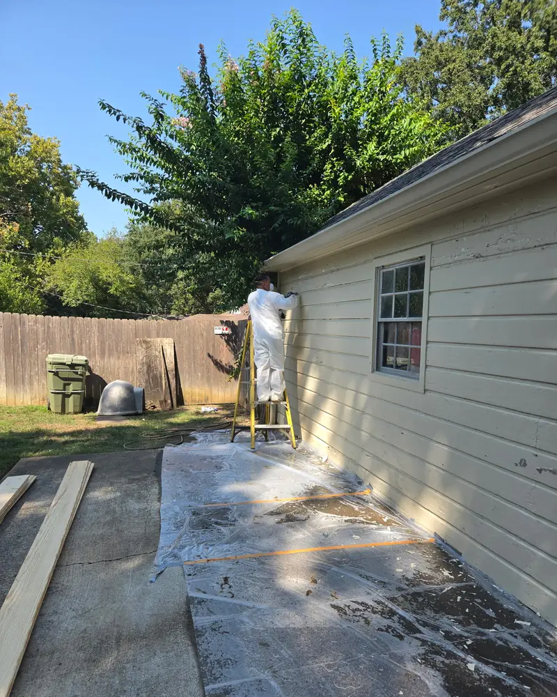 A worker in a white suit and mask on a ladder at the roof edge of a cream wood-sided house, with plastic sheeting on the driveway and new unpainted boards lying to the left