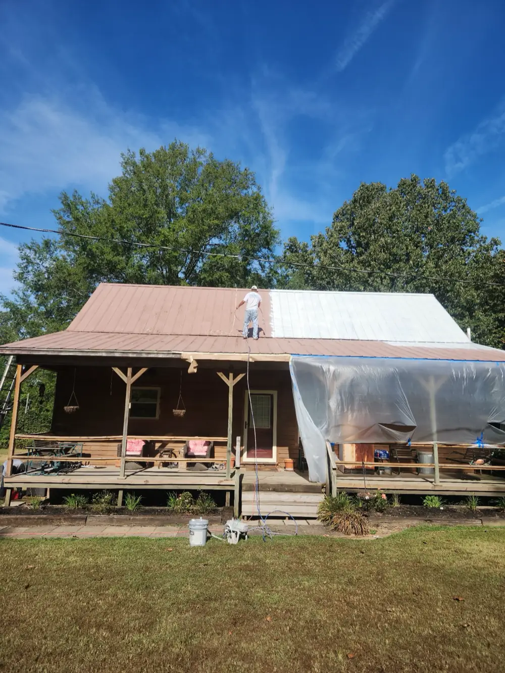 A metal roof half sprayed white over the original rust red, with the porch below sheeted in plastic